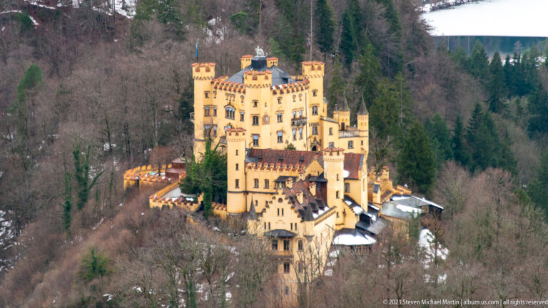 Ochre Hohenschwangau Castle with crenellated towers and red roofs standing above a wooded winter hillside.