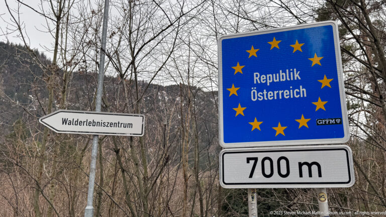 Blue “Republik Österreich” border sign with EU stars, “700 m” plaque, and Walderlebniszentrum direction sign.