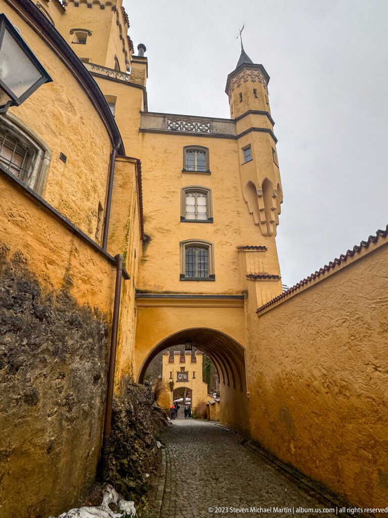 Cobbled lane passing beneath an ochre arched passage within Hohenschwangau Castle, with towers and crenellated walls above.