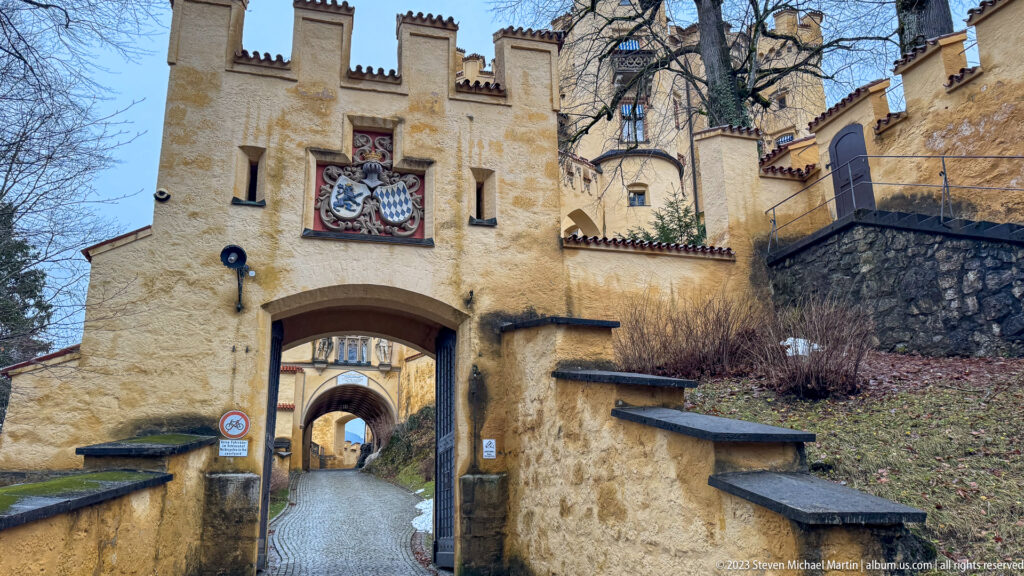 Ochre crenellated gateway with a painted heraldic panel above a round arch at Hohenschwangau Castle.
