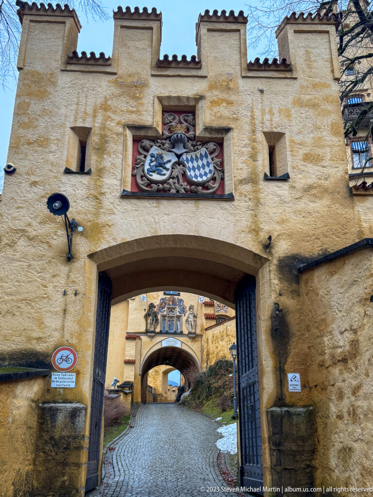 Close frontal view of Hohenschwangau Castle's ochre gateway, painted coat of arms, crenellations, and open timber gates.
