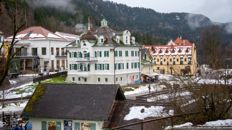 White Jägerhaus with green shutters among snowy buildings at the foot of a wooded mountain slope.