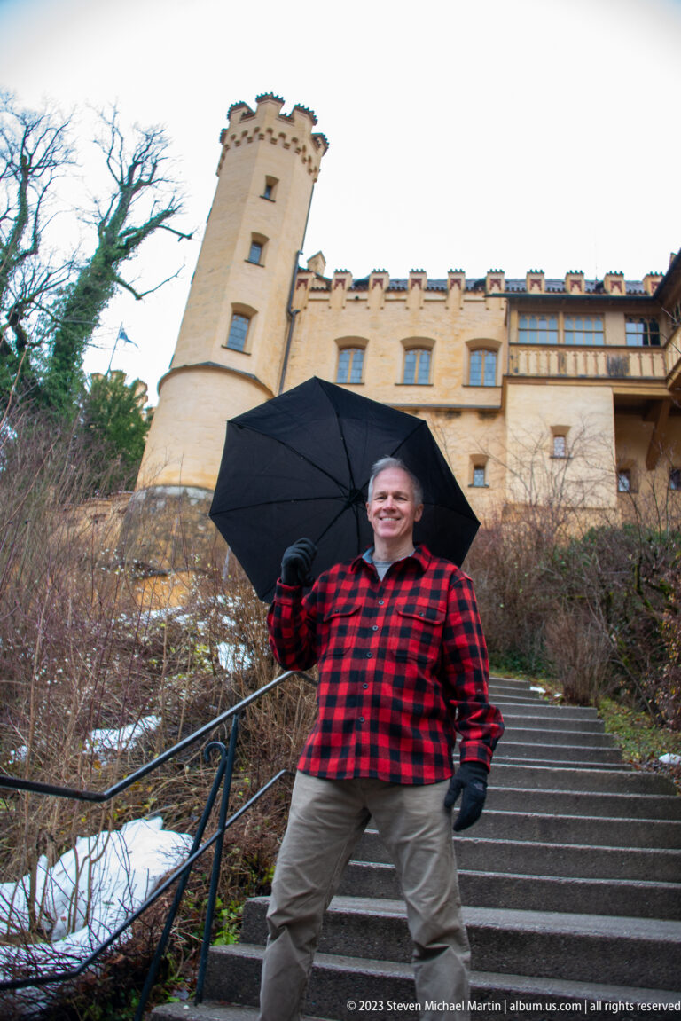 Male visitor holding a black umbrella on stone steps below the ochre walls and tower of Hohenschwangau Castle.
