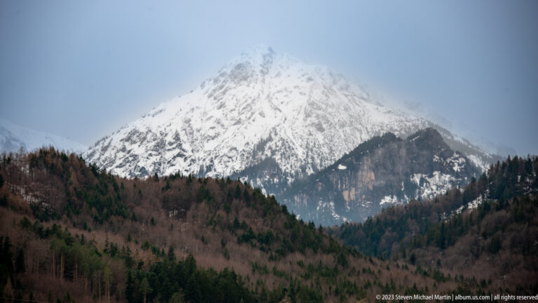 Snow-covered Alpine mountain rises above dark forest, with its summit softened by cloud.