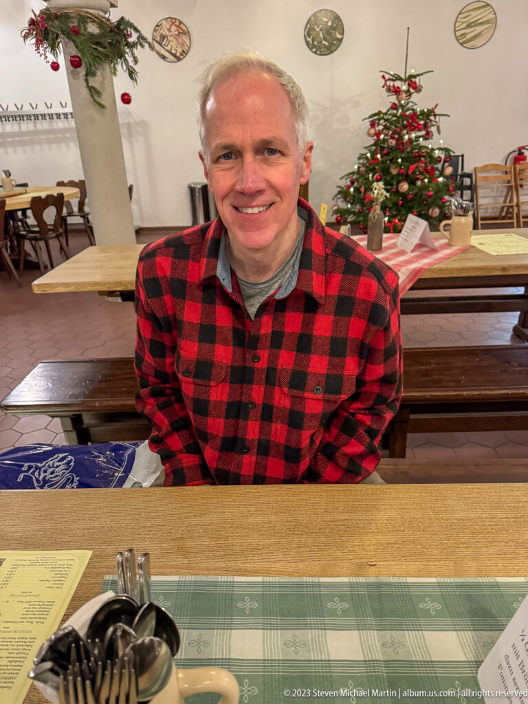 Male visitor in a red checked jacket seated at a dining table with a decorated Christmas tree behind him.