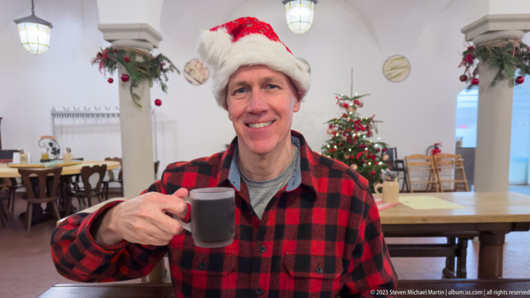 Male visitor in a Santa hat raising a mug of dark hot drink beside a decorated Christmas tree.