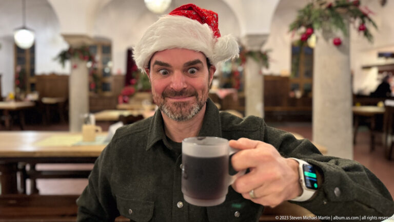 Male visitor smiling broadly and raising a mug of dark drink inside a Christmas-decorated restaurant.