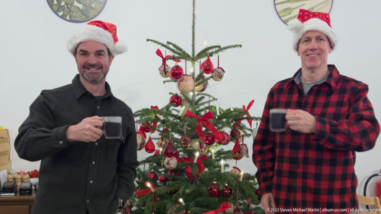 Two male visitors holding mugs beside a Christmas tree decorated with red ribbons and ornaments.