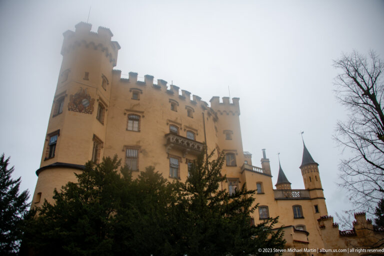 Ochre Hohenschwangau Castle rising above trees, with crenellations, balcony, towers, and misty sky.