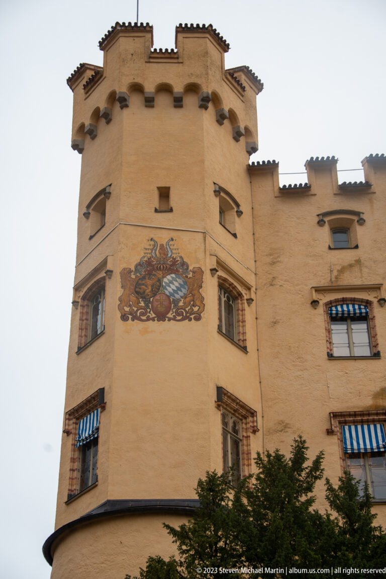 Painted heraldic arms on the ochre tower of Hohenschwangau Castle beneath crenellations and shuttered windows.