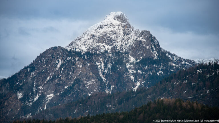 Craggy pyramidal Alpine summit streaked with snow above forested lower slopes.