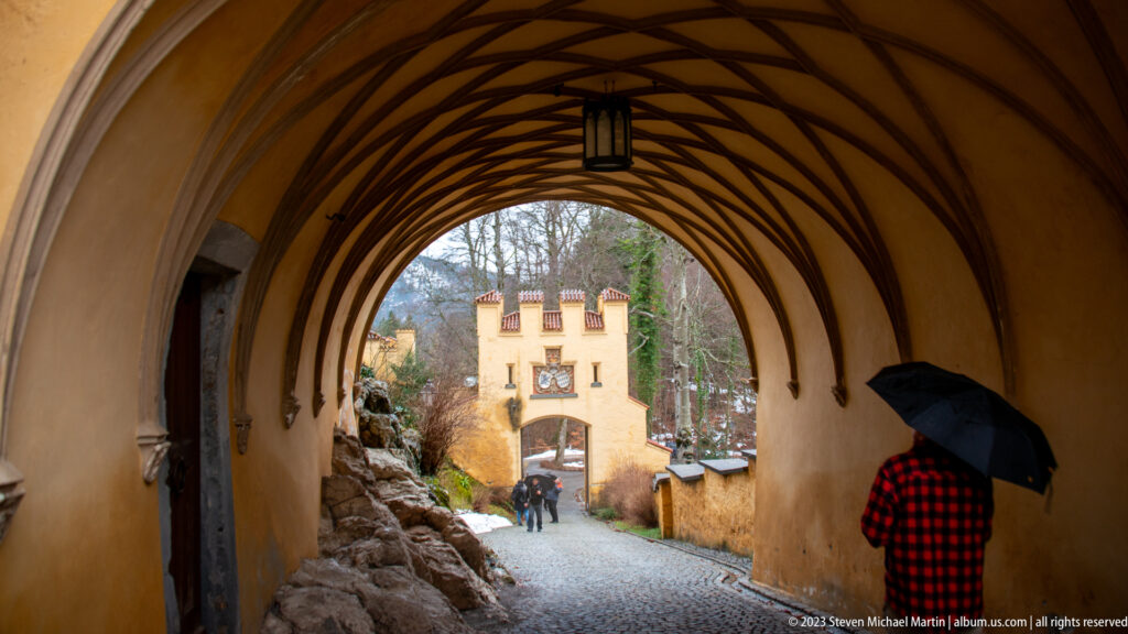 Ribbed vaulted passage leading toward an ochre crenellated gate, with a visitor holding an umbrella at right.