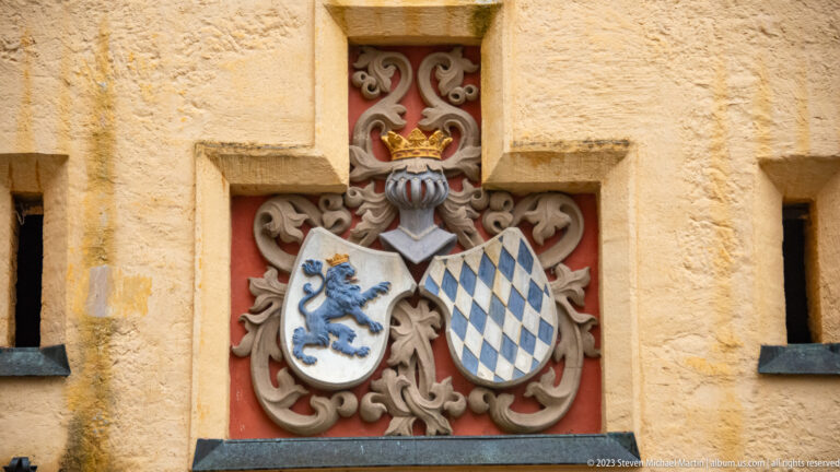 Carved heraldic relief with crowned helmet, rampant lion shield, and blue-and-white Bavarian lozenges.