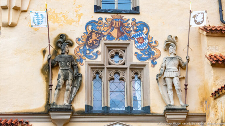 Two armored statues flanking a narrow tracery window beneath blue-and-gold heraldic painting on Hohenschwangau Castle.