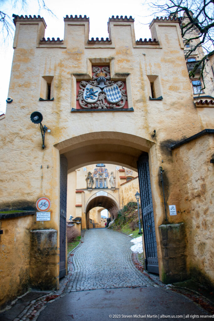 Broad ochre arched gate with heraldic panel and a second gateway visible beyond along a cobbled road.