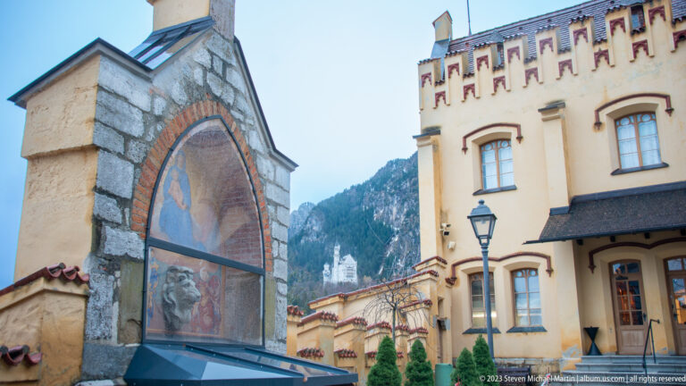 Painted pointed wall panel beside ochre Hohenschwangau Castle buildings, with mountains and distant Neuschwanstein visible.