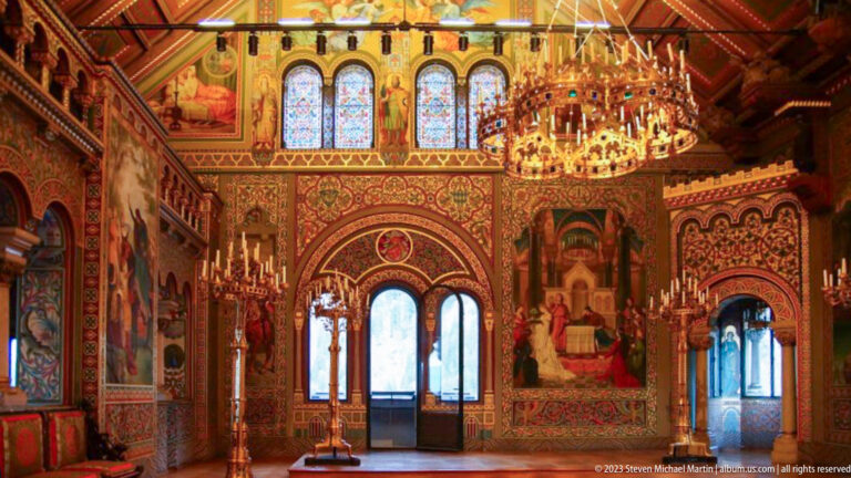 Richly painted Hohenschwangau Castle interior with murals, stained glass, arched doorways, and a large chandelier.