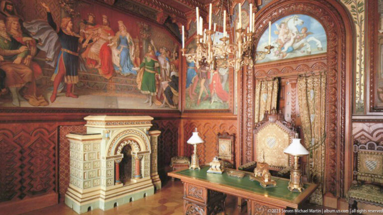 Furnished Hohenschwangau Castle room with wall murals, tiled stove, writing desk, lamps, and carved wood furniture.