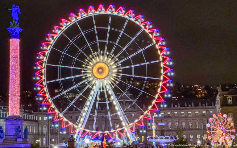 Illuminated Ferris wheel beside the Jubilee Column on Stuttgart Schlossplatz at night, with historic buildings and Christmas lights behind.