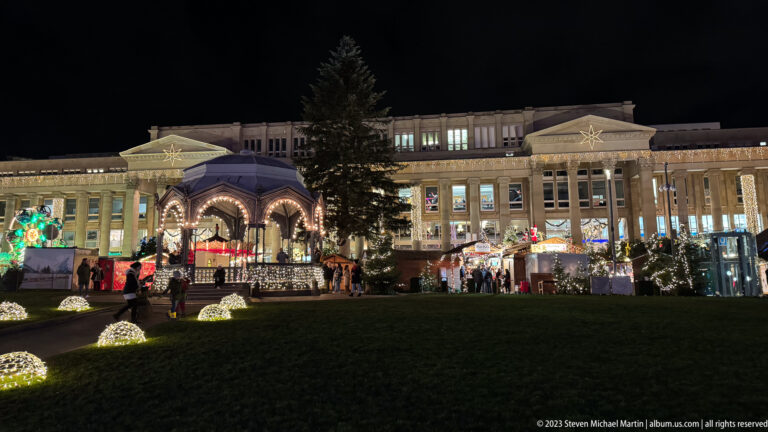 Tall Christmas tree, illuminated pavilion, glowing lawn ornaments, and the Königsbau across Stuttgart Schlossplatz at night.