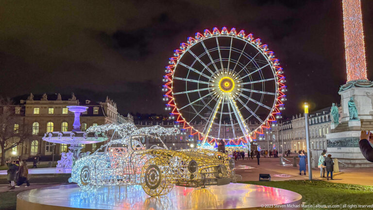 White illuminated wireframe Mercedes-Benz roadster sculpture on Schlossplatz with a Ferris wheel glowing behind it.