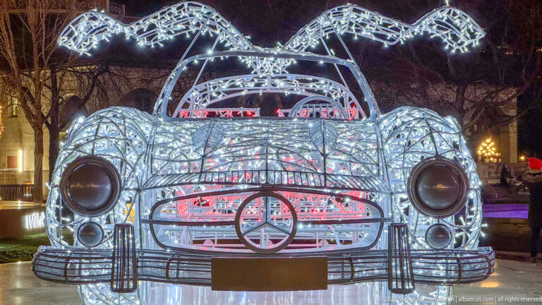 Cool-white illuminated Mercedes-Benz roadster sculpture viewed head-on, with wireframe fenders, headlights, grille, and open cockpit.