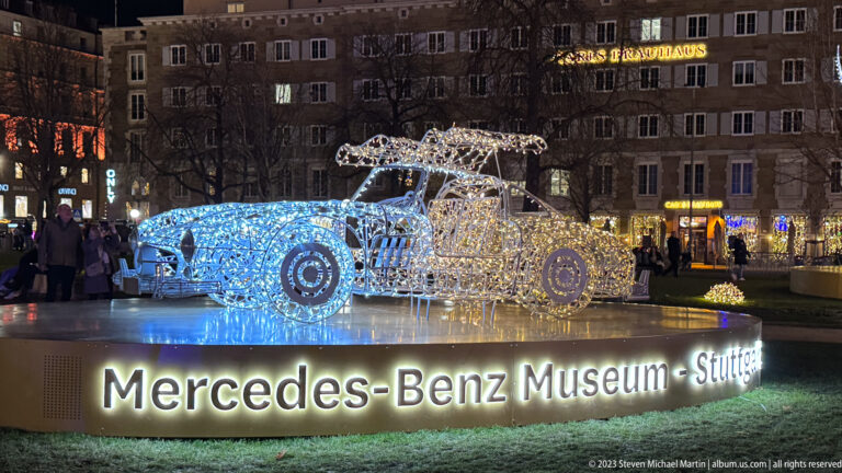 Two illuminated vehicle sculptures above a glowing “Mercedes-Benz Museum – Stuttgart” sign, with pedestrians and buildings behind.