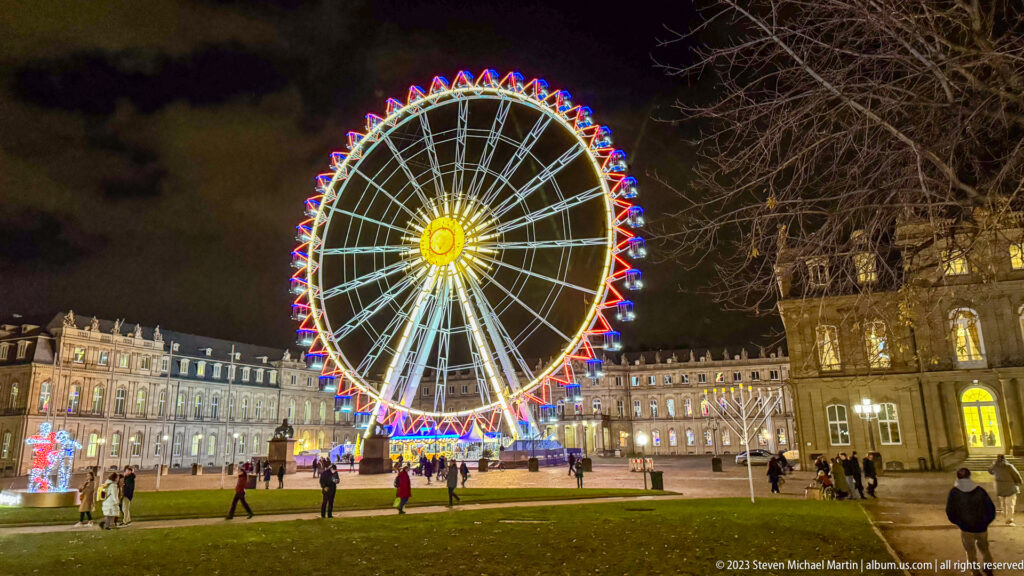 Wide nighttime view across Schlossplatz lawn toward an illuminated Ferris wheel and historic palace façades, with scattered visitors.