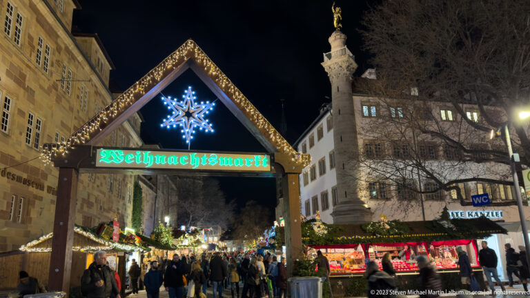 Lit timber gable reading “Weihnachtsmarkt” above a busy market entrance, with stalls, visitors, and a tall stone column beyond.