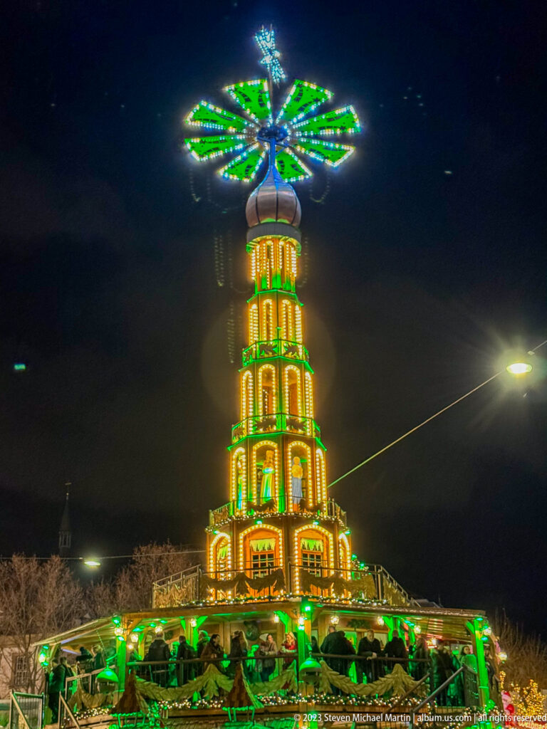 Large green-and-gold tiered Christmas pyramid with a rotating fan above a crowded Stuttgart market stall at night.