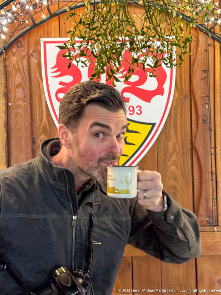 Man sipping from a small mug beneath evergreen branches and a large VfB Stuttgart crest on a wooden market stall.