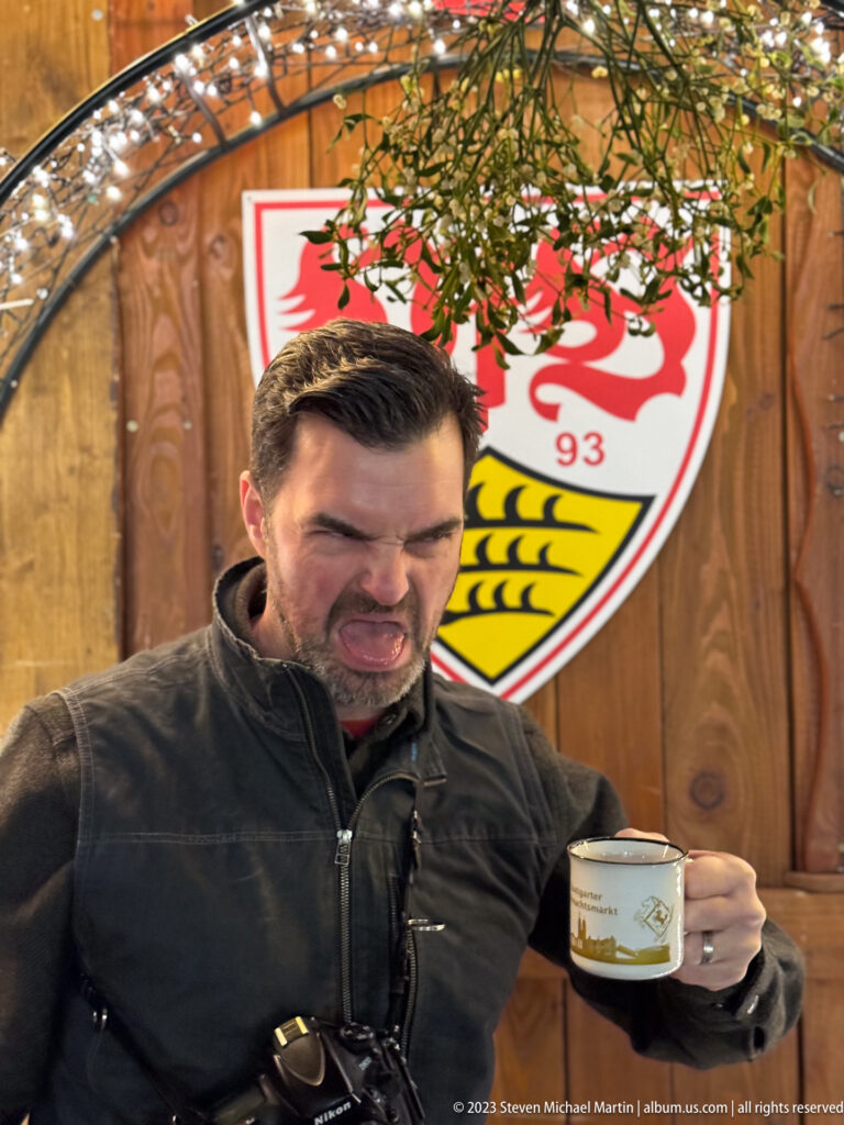 Man holding a small mug and making a playful face beneath evergreen branches and a VfB Stuttgart crest.