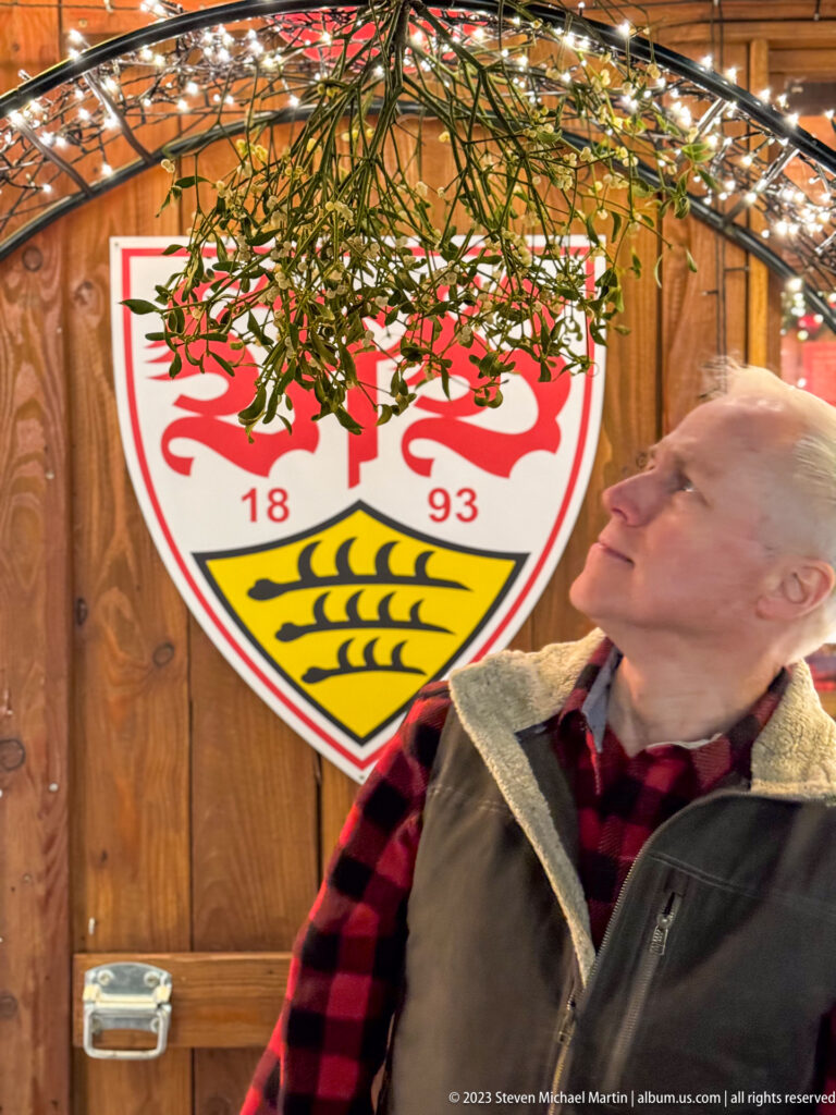 Older man in a red-and-black checked shirt looking upward beneath greenery, with a large VfB Stuttgart crest behind him.