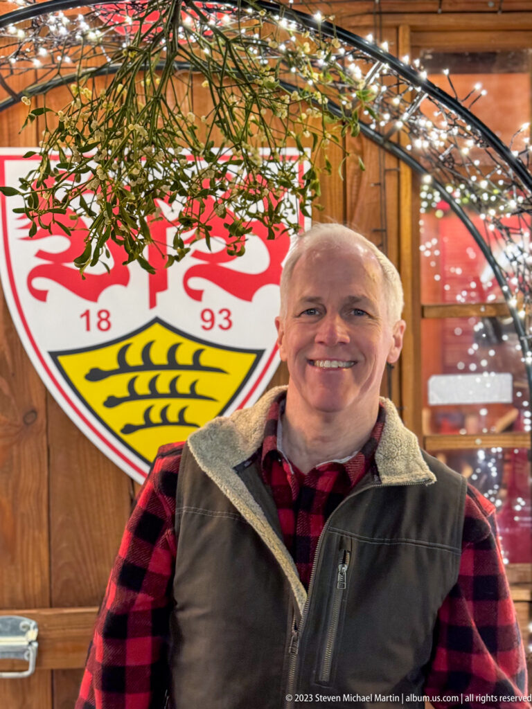 Smiling older man facing the camera beneath greenery and a large VfB Stuttgart crest on a wooden market stall.
