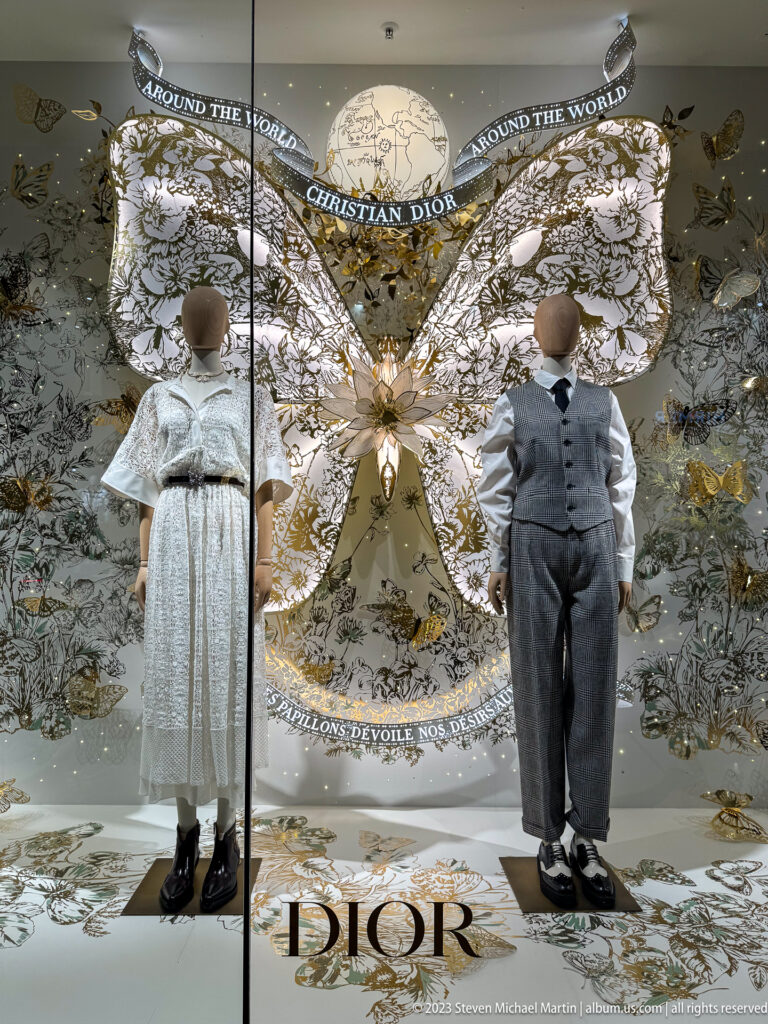 Two child mannequins in pale formal clothing before an ornate butterfly-shaped Dior window display with black-and-white botanical graphics.