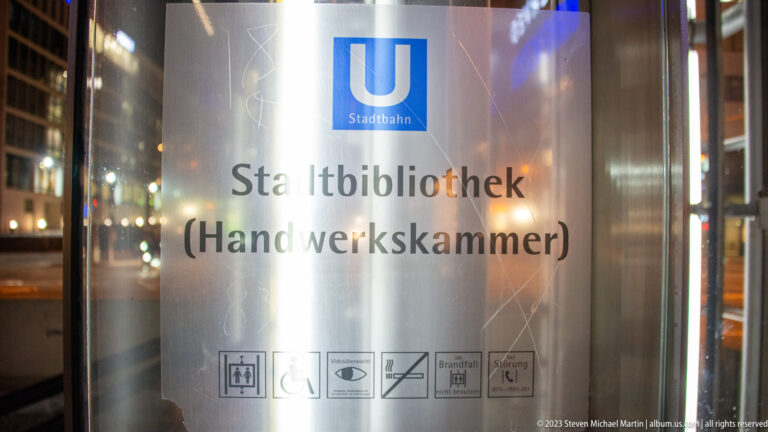Glass transit panel with blue U-Bahn symbol and the words “Stadtbibliothek (Handwerkskammer),” plus accessibility and safety pictograms.