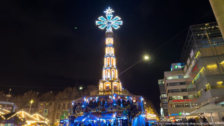 Tall amber-lit Christmas pyramid with a glowing rotor, framed by modern city buildings and overhead wires at night.