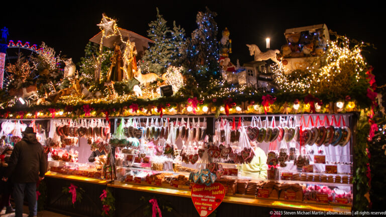 Night view of an illuminated Christmas-market sausage stall with hanging meats and a rooftop scene of miniature houses, trees, figures, and animals.