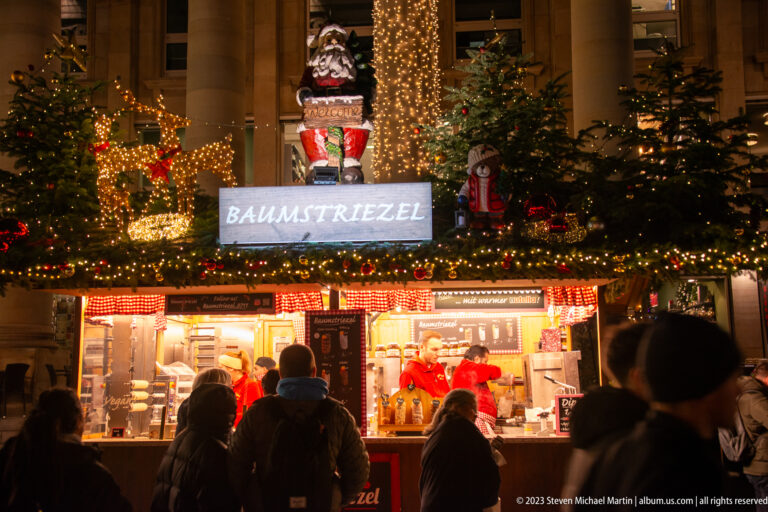 Customers stand at a brightly lit Baumstriezel stall decorated with fir trees, nutcrackers, wreaths, and Christmas lights.