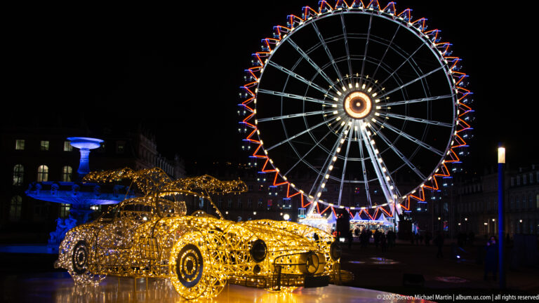 Golden car-shaped light sculpture in front of a large illuminated Ferris wheel on Stuttgart’s Schlossplatz at night.