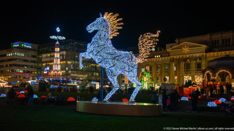 Large illuminated rearing horse sculpture on a circular base amid Christmas lights and crowds in central Stuttgart.