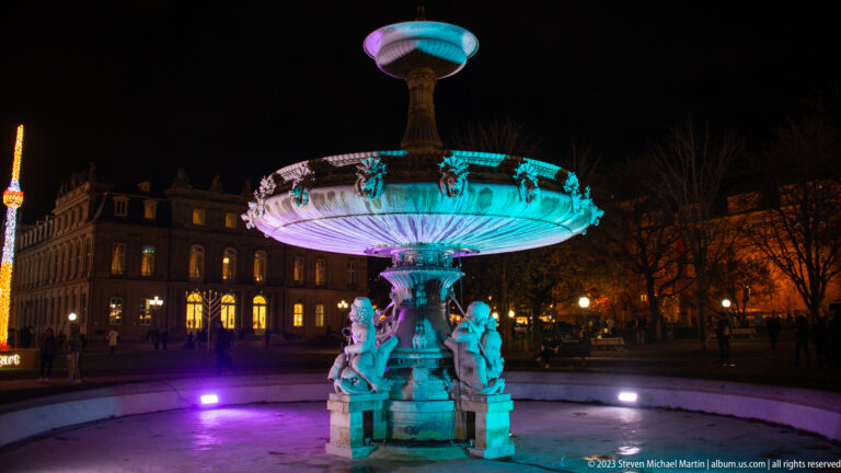 Ornate sculptural fountain lit turquoise and violet at night, with historic buildings and winter trees behind.