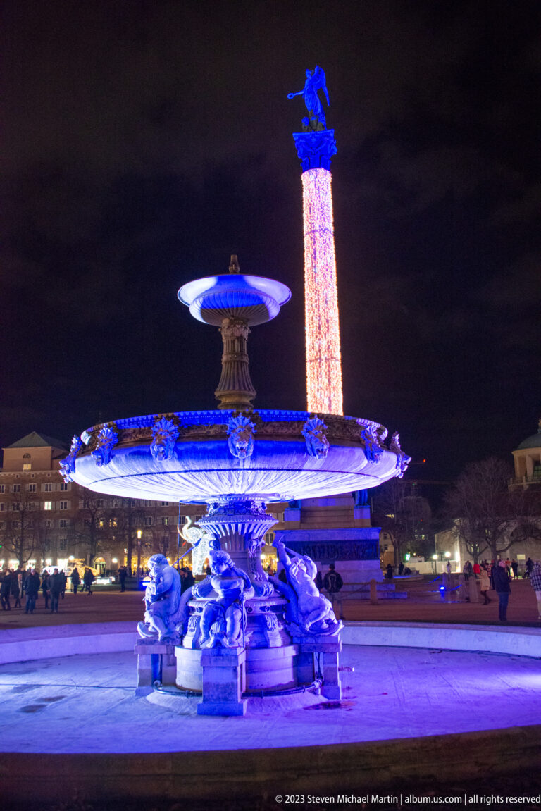 Blue-lit sculptural fountain in the foreground with Stuttgart’s tall Jubiläumssäule rising behind at night.