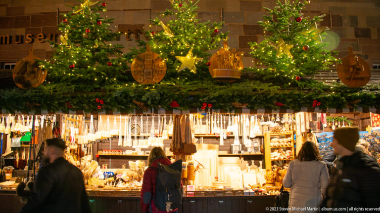 Shoppers browse a Christmas market stall filled with wooden ornaments and crafts beneath decorated fir trees.