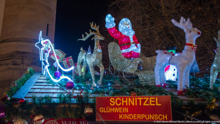 Santa, reindeer, sleigh, and snowman decorations above a stall sign for schnitzel, Glühwein, and Kinderpunsch.