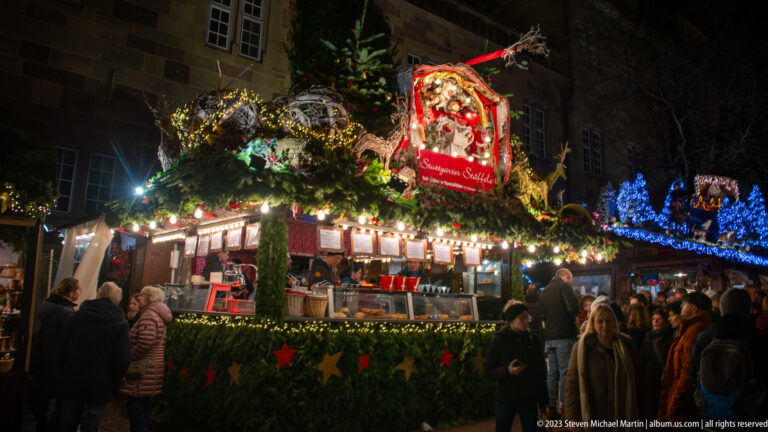 Crowded Stuttgart Christmas market stall with garlands, lights, oversized ornaments, and visitors gathered around the counter.