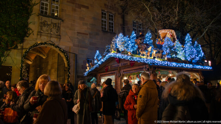 Blue-lit fir trees and woodland figures top a crowded Christmas market stall beside a stone arched doorway.