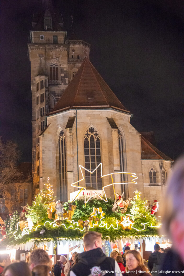 Stone choir and tiled roof of Stuttgart’s Stiftskirche rising behind a brightly decorated Christmas market stall.