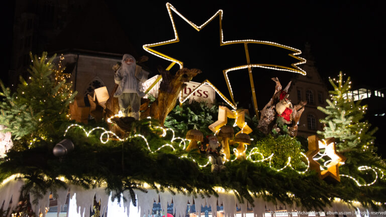 Large illuminated shooting star above Santa figures, animals, gifts, and fir branches on a Christmas market stall roof.