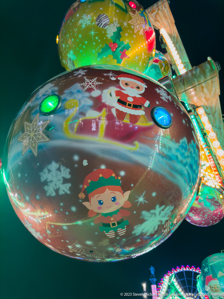 Close-up of a curved amusement-ride panel painted with an elf, Santa figure, snowflakes, ornaments, and colored bulbs.